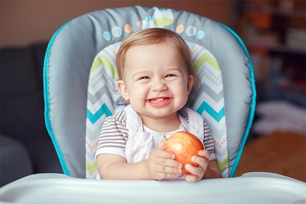 A girl, sitting on a high chair, biting on an apple.