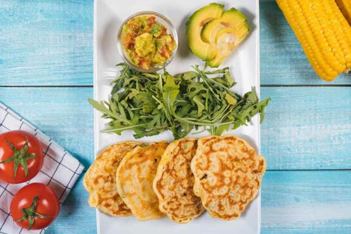 A plate of avocado corn fritters with herbs, guacamole, and avocado slices on the side. Two tomatoes, napkins, and corn cob lie on the table around the plate.